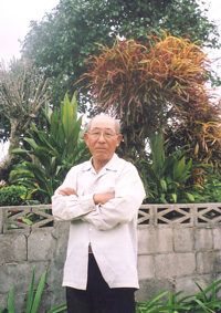 親木の前に立つ南風原さん。種から1本1本植えたツバキは20年かけて数万本に